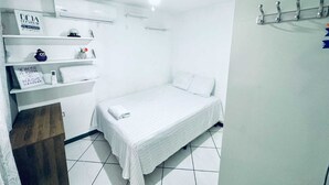 A small, white, single-bed room with a ceiling fan, a wall-mounted shelf with toiletries, and a bed with a white comforter and pillows.