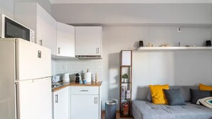 A compact kitchen with white cabinets, a built-in oven, and a coffee machine. There's a grey sofa with yellow and grey cushions in the living area. - Athenian Entertainment & Relaxation Hub (Athens)