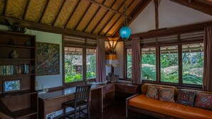 A room with a wooden desk, a chair, a sofa with patterned cushions, a bookshelf, and a large window with a view of greenery.