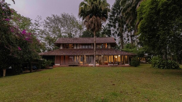 Una casa de dos pisos con techo de tejas, rodeada de abundante follaje y árboles.