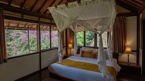 A room with a bed, a canopy, a nightstand, and a window with a view of purple flowers.