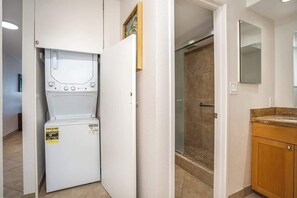 Une buanderie équipée d’un lave-linge et d’un sèche-linge à chargement frontal, d’une douche avec une porte en verre et d’un meuble-lavabo en bois avec un miroir.