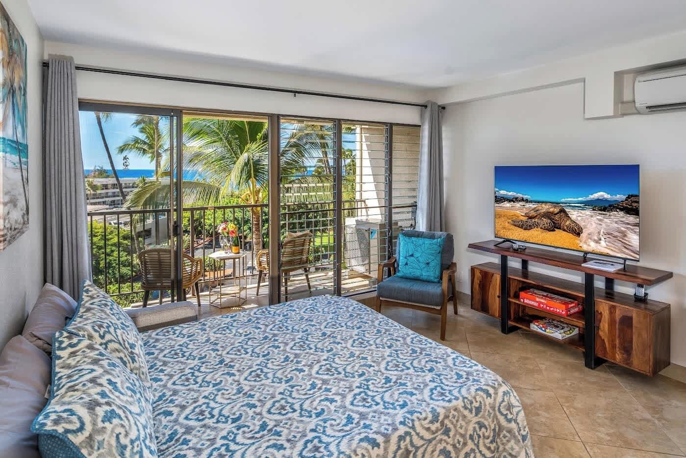 A hotel room with a large bed, a TV, a balcony with a view of the ocean, and a dining area with chairs and a table.