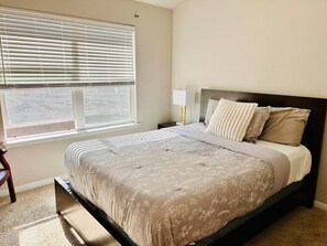 Un dormitorio con cama, mesita de noche, lámpara y ventana con persianas.