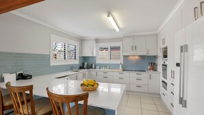A modern kitchen with white cabinets, a central island with a bowl of fruit, and a dining area with two wooden chairs. - Bren Place, 6, Battery Hill (Battery Hill)