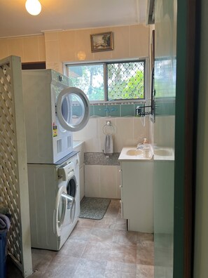 A compact laundry room with a washing machine, dryer, and a small sink with a window above. - XYL guests house (Caloundra)
