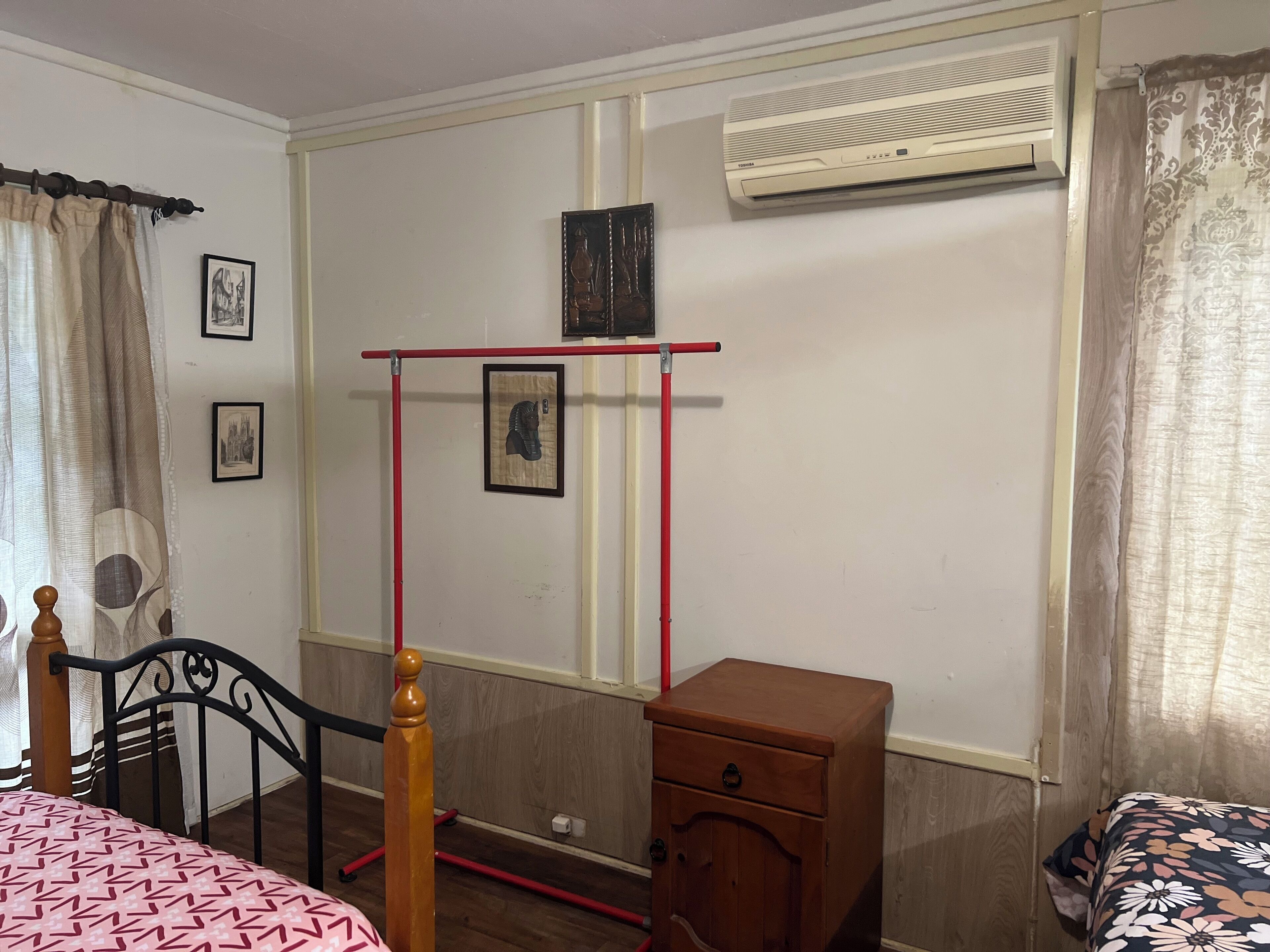 A bedroom with a bed, a wooden nightstand, a red and black metal rack, an air conditioner, and framed pictures on the wall.