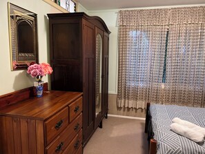 A bedroom with a wooden dresser, a mirror, a vase with flowers, a bed with a patterned bedspread, and a window with floral curtains. - XYL guests house (Caloundra)