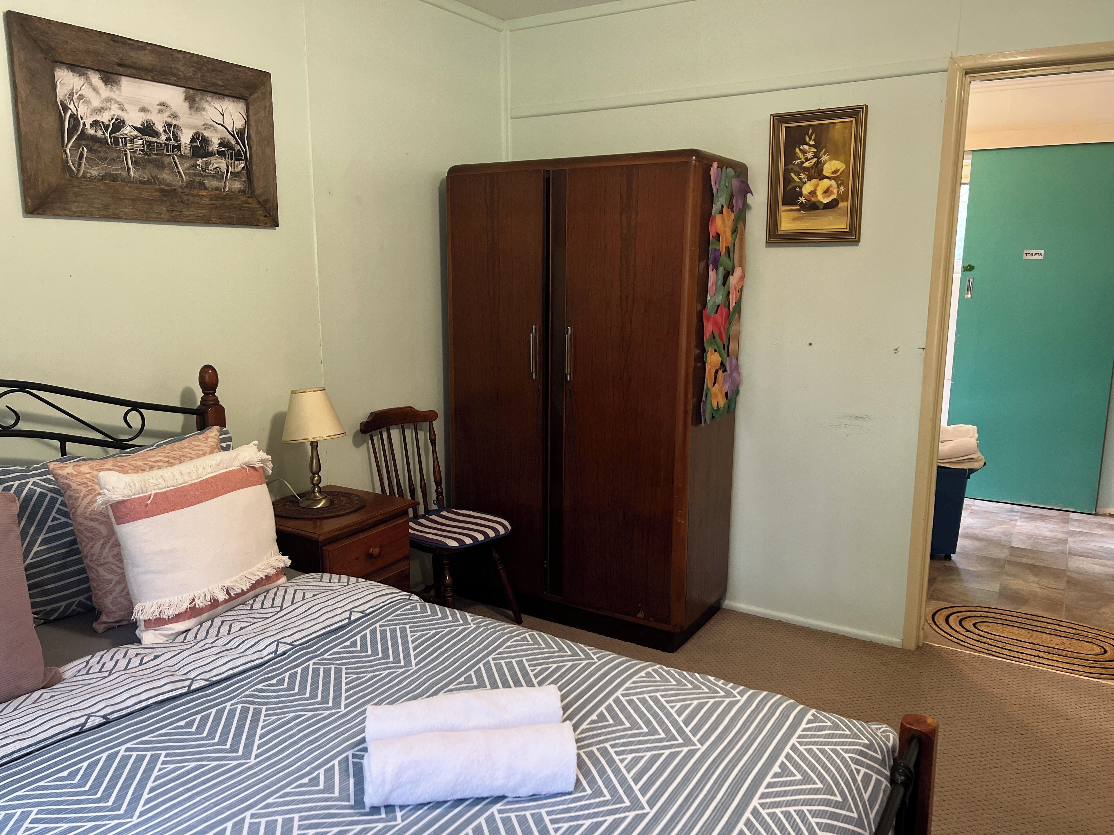 A bedroom with a bed, a wooden wardrobe, a chair, a desk with a lamp, and a picture on the wall.