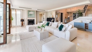 Un salon spacieux avec un grand canapé blanc, une table basse et une cheminée. Un escalier mène à un niveau supérieur, et on y trouve un meuble bleu orné d’objets décoratifs.