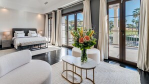 Une chambre d’hôtel moderne dotée d’un grand lit, d’un coin salon avec une table basse et offrant une vue sur une piscine et des palmiers.