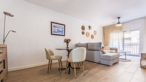 Un salon moderne avec un canapé gris, deux fauteuils, une petite table ronde et une table à manger. Un tableau encadré orne le mur, un ventilateur de plafond est installé au plafond, et une porte coulissante offre une vue sur l’extérieur.