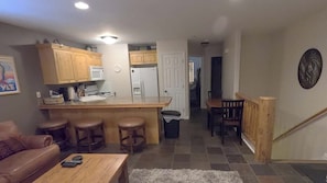 Cucina con mobili in legno, forno a microonde e frigorifero. È presente una zona bar con sgabelli e un tavolino. Si intravede un cestino della spazzatura vicino alla cucina.