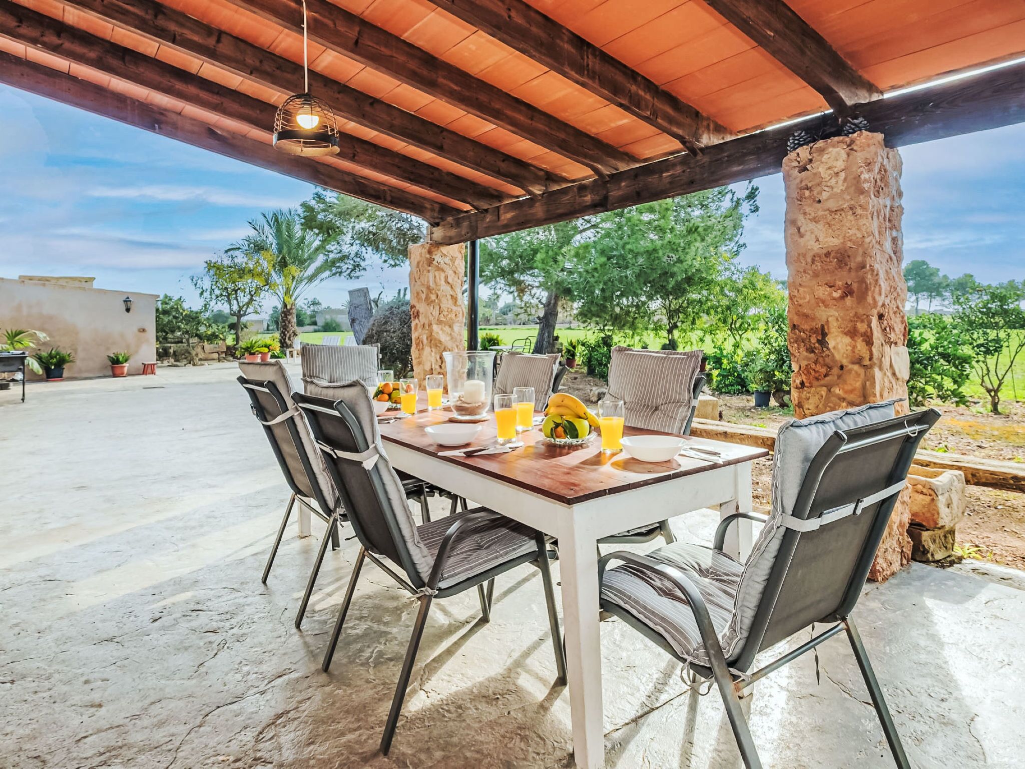 Um pátio externo com mesa de madeira posta para refeições, cercado por cadeiras.
