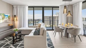 A modern living room with a sofa, dining table, and chairs, large windows with a city view, and a decorative vase on a side table.
