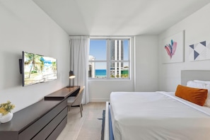 Habitación de hotel con cama, escritorio, televisor y ventana con vistas a edificios y al mar.