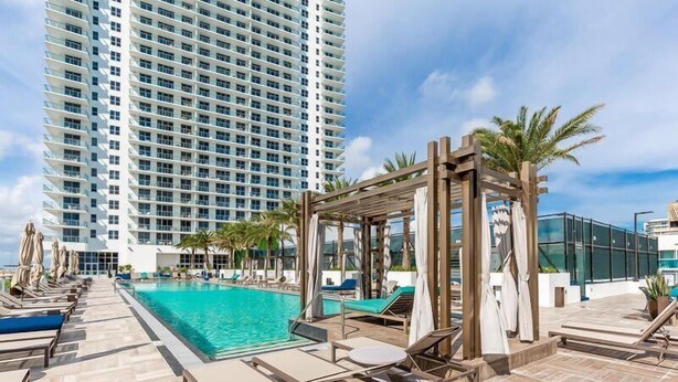 A hotel pool area with a clear view of a tall building in the background, lounge chairs, and a wooden pergola structure.