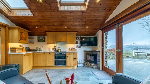 A modern kitchen with wooden cabinets and a stone fireplace, a living area with leather sofas, and a large window offering a view of the countryside. - Stoney Calf -  Quaint Cottage for two on a working farm (Old Town)