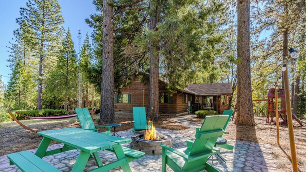 A wooden cabin surrounded by trees, a fire pit, and green picnic tables and chairs.