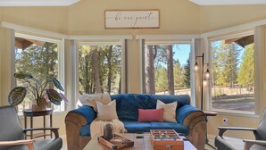 A cozy living room with a blue sofa, a coffee table with a foosball table, and a large window offering a view of trees.