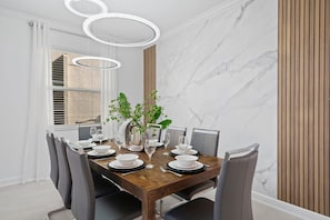 Uma sala de jantar com uma mesa de madeira posta para seis pessoas, pratos brancos e um vaso com plantas verdes.