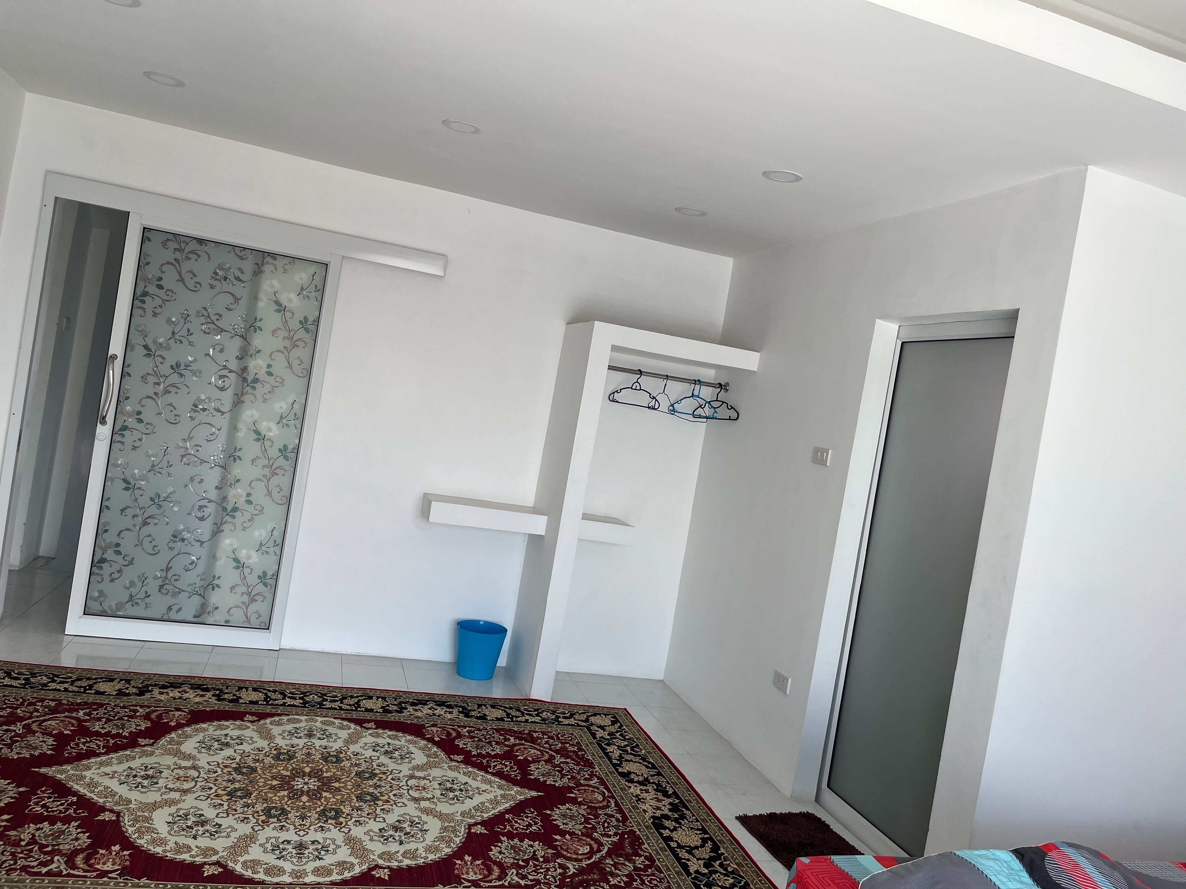 A room with a patterned carpet, a sliding door with floral curtains, a blue trash bin, and a coat rack.