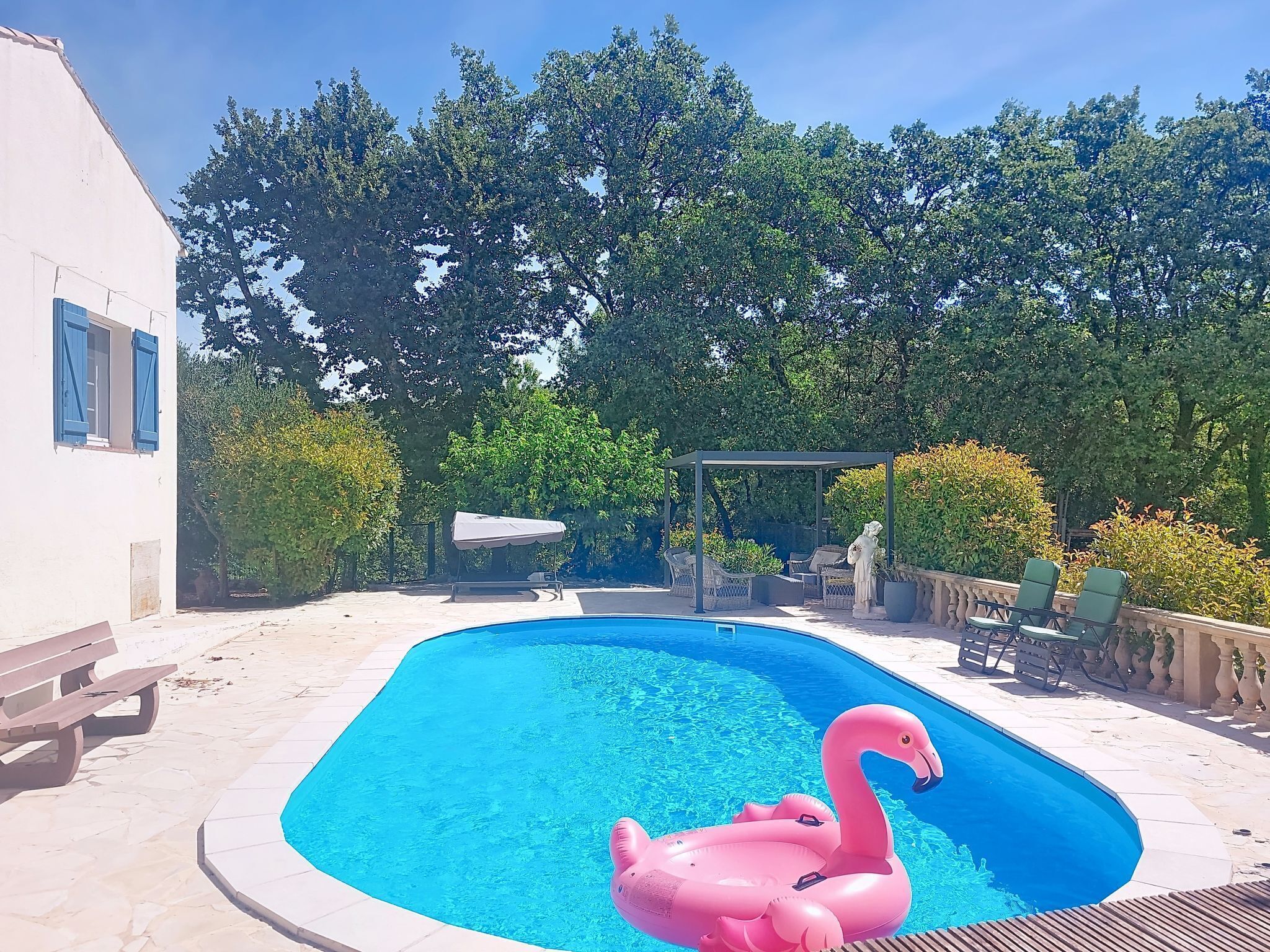 Zwembad met een roze flamingo drijver, omgeven door een wit gebouw, bomen en tuinmeubilair.