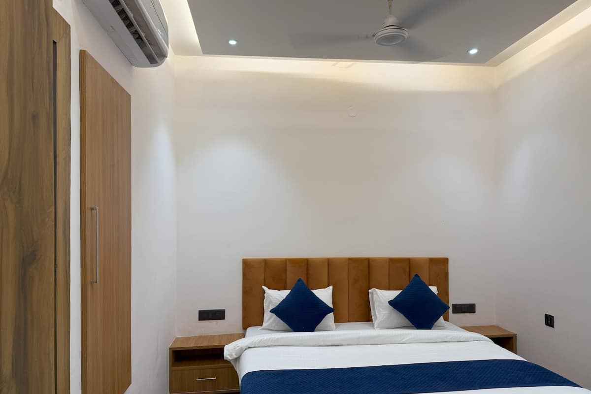 Une chambre d’hôtel avec une tête de lit en bois, un lit avec une literie blanche et des touches de bleu, une table de chevet en bois et un climatiseur fixé au mur.