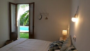 A bedroom with a bed, a bedside table with a lamp, a wall clock, and a view of a pool through an open door. - Son Floriany - House with private pool and lounge (Son Severa)