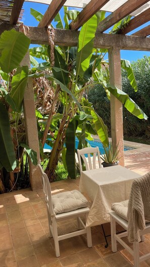 Un espacio al aire libre cubierto con mesa y sillas, rodeado de plataneros y vistas a una piscina.