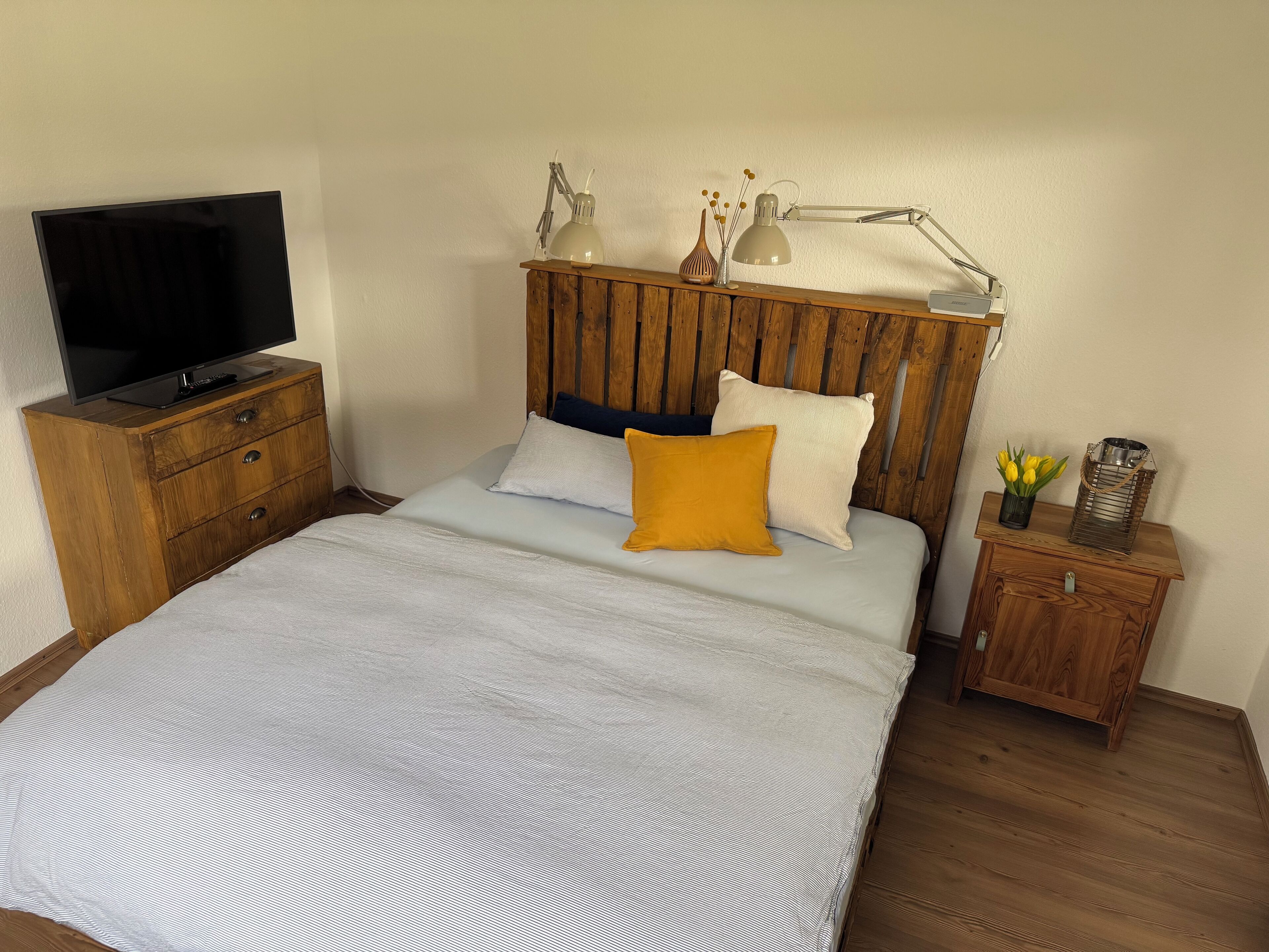 Ein Schlafzimmer mit einem hölzernen Kopfende, einem Bett mit gestreifter Bettdecke, einem Nachttisch mit Lampe, einem Fernseher auf einer Holzkommode und einem kleinen Tisch mit Blumen in einer Vase.