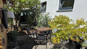 Ein Sitzbereich im Freien mit Tisch und Stühlen, umgeben von Topfpflanzen und einem Gebäude im Hintergrund.
