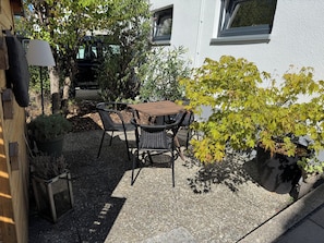 Un espace extérieur aménagé avec une table et des chaises, entouré de plantes en pot et avec un bâtiment en arrière-plan.