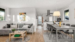 Un salon moderne avec un canapé gris, une table basse et un coin repas comprenant une table et des chaises. On aperçoit une cuisine à l’arrière-plan.
