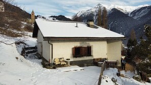 A small, snow-covered house with a wooden porch and a stone wall. - Chasa Patgific: Cozy 3-Room Holiday Apartment with Wood Stove and Stunning Garden in Ramosch (Ramosch)