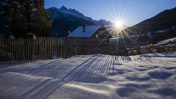 Paesaggio innevato con una recinzione di legno, una casa dal tetto spiovente e montagne sullo sfondo.