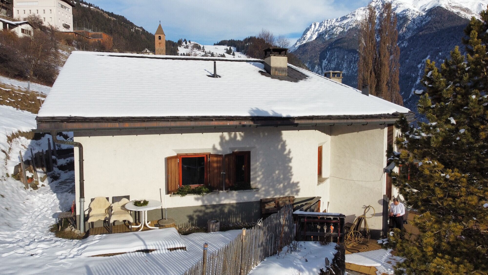 A snow-covered house with a small porch, a wooden window, and a view of snow-capped mountains in the background.