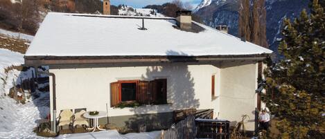 A snow-covered house with a small porch, a wooden window, and a view of snow-capped mountains in the background.