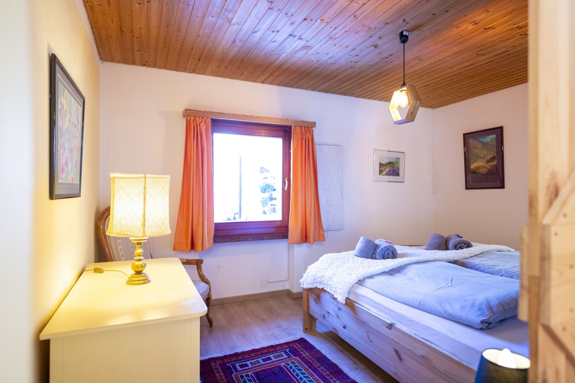 Ein Schlafzimmer mit Holzbalkendecke, einem Bett mit weißer Decke, einem Schreibtisch mit Lampe und einem Fenster mit orangefarbenen Vorhängen.