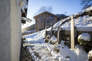 A snow-covered outdoor area with a wooden house, a stone wall, and a wooden fence. - Chasa Patgific: Cozy 3-Room Holiday Apartment with Wood Stove and Stunning Garden in Ramosch (Ramosch)