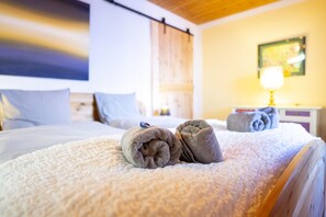 Una cama bien hecha con toallas enrolladas, cabecera de madera, lámpara de noche y un cuadro en la pared.