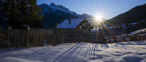 Schneebedeckte Landschaft mit Holzzaun, Haus mit geneigtem Dach und Bergen im Hintergrund.
