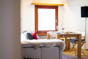A cozy room with a bed, a dining table set for two, and a window with a view. - Chasa Patgific: Cozy 3-Room Holiday Apartment with Wood Stove and Stunning Garden in Ramosch (Ramosch)