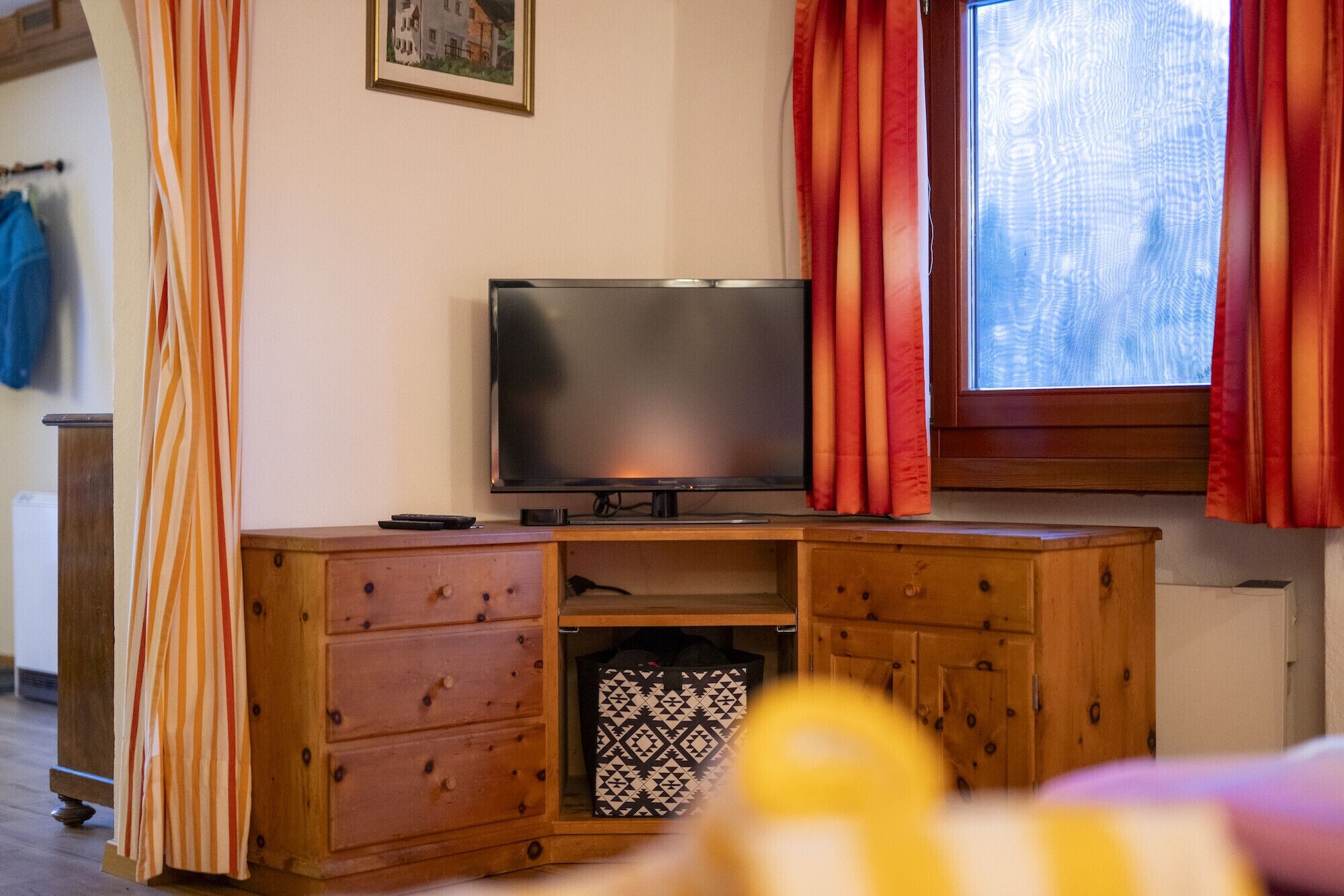 Ein Wohnzimmer mit einem Fernseher auf einer Holzkassette, einem Fenster mit roten Vorhängen und einer dekorativen Aufbewahrungsbox.