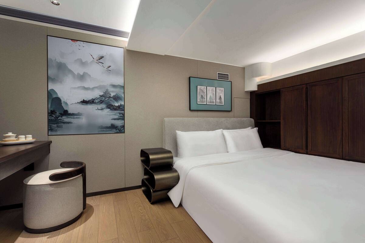 Ett modernt hotellrum med en stor säng, ett skrivbord och en papperskorg. Det finns en tavla på väggen och en inramad bild ovanför sängen.