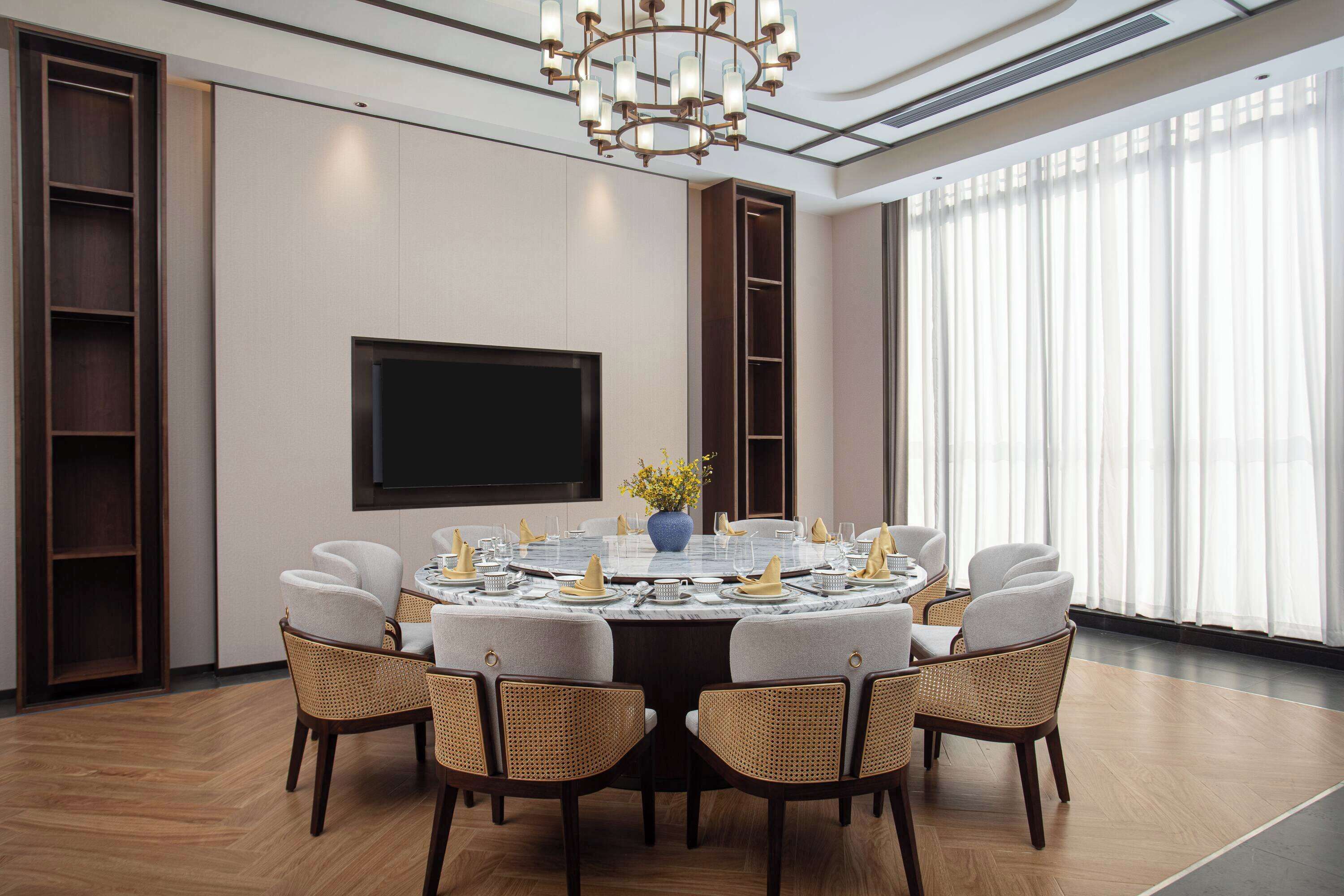 Un comedor moderno con una mesa redonda, seis sillas, un televisor de pantalla plana y un amplio ventanal con cortinas translúcidas.