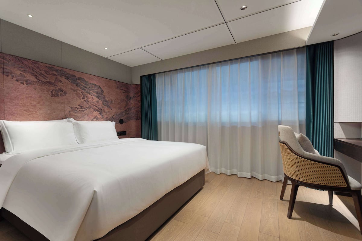 Ett modernt hotellrum med en stor säng, en sittgrupp med en fåtölj och utsikt över omgivningarna genom tunna gardiner.