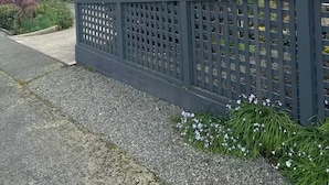 A two-story house with a grey exterior, a red-framed window, and a small garden with pink flowers and a wooden fence. - Heart of Windsor (Invercargill)
