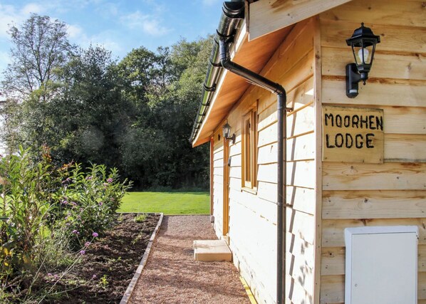 Una construcción de madera con el cartel 'MOORHEN LODGE', con un camino que lleva a la entrada, rodeada de follaje.