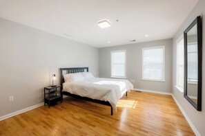 A bedroom with a wooden floor, a bed with white bedding, a nightstand with a lamp, and two windows with blinds.